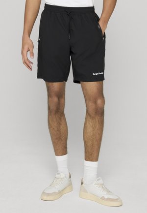 Hombre con pantalones cortos negros de Sergio Tacchini, calcetines blancos y zapatillas blancas, de pie con una mano en el bolsillo frente a un fondo liso.