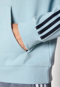 Light blue sweatshirt with ribbed cuffs and hem; features navy three-stripe design along the arm and a front pocket.
