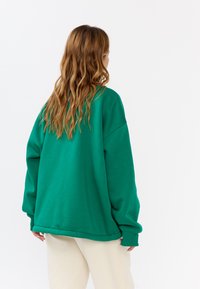 Felpa verde oversize realizzata in tessuto morbido, con maniche ampie e scollo rotondo. La parte posteriore ha una texture liscia e una vestibilità comoda.