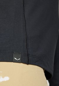 Crna majica dugih rukava od tkanine s teksturom. Sadrži detalj šava i mali naziv s bijelim logotipom na donjem lijevom rubu.
