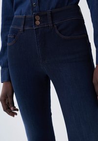Jeans de mezclilla de color azul oscuro con una cintura alta, cierre de doble botón y detalles de costura naranja. Presenta bolsillos frontales y un ajuste slim.