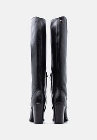 Ζευγάρι ψηλών μαύρων δερμάτινων μπότες με χοντρά τακούνια, από πίσω, σε απλό λευκό φόντο.