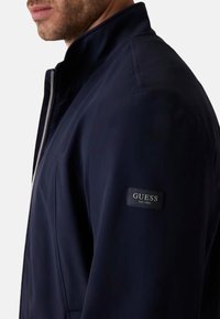 Giacca navy con colletto rialzato, chiusura con zip e tessuto testurizzato. Presenta una patch con il marchio sulla manica sinistra.