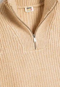 Jersey de punto beige con cuello de media cremallera. Tejido suave y texturizado con detalle de canalé visible, cuenta con una pequeña etiqueta colgante para el tamaño.