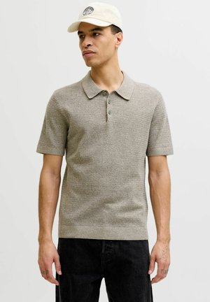 Férfi bézs színű kötött rövid ujjú galléros pólót és világos színű sapkát visel, sima háttér előtt áll.