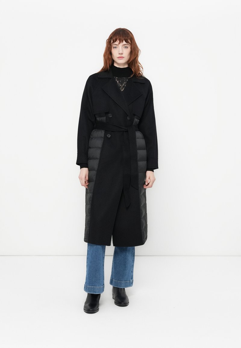 Lungo cappotto nero con un design doppiopetto, dotato di imbottitura trapuntata sui lati, una cintura e un colletto. Abbinato a jeans svasati e stivaletti al polpaccio.