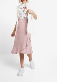 Bílý blejzr, květinové tričko s grafikou a midi sukně s pruhy v červené a bílé barvě se volánovým detailem, doplněné bílými teniskami.