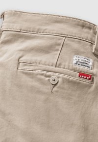 Poche de pantalon beige Levi's avec fermeture par bouton, onglet rouge Levi's visible et étiquette blanche de la marque au-dessus de la poche.