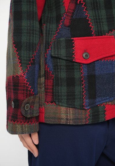 Καρό σακάκι με πατς, σε πράσινο, κόκκινο και μπλε χρώμα, με κόκκινη ραφή και μανσέτες με κουμπιά. Διαθέτει εμπρόσθια τσέπη.