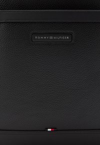 Čierna kožená taška s textúrovaným povrchom, s horizontálnym popruhom a striebornou logovou tabuľkou. Doplněná malým trojfarebným detailom na spodnej časti.