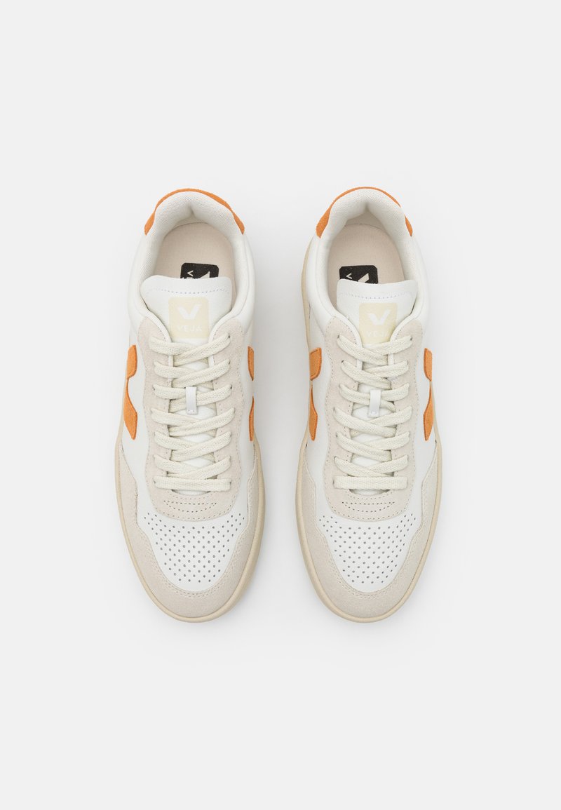Baskets de sport blanches avec des accents orange, présentant une boîte à orteils perforée, un design à lacets et des panneaux latéraux en daim beige texturé.