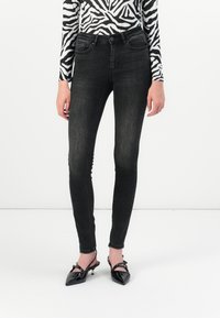 Vaqueros ajustados de mezclilla negra con una cintura alta, un ligero desgaste, cinco bolsillos y detalles de costura, combinados con zapatos de punta negra de charol.