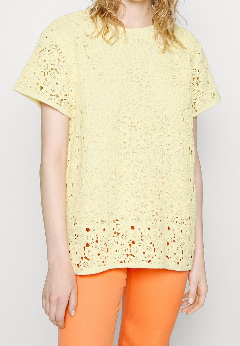 Haut en dentelle jaune pâle, avec un motif floral, manches courtes et ourlet festonné, associé à un pantalon orange vif.