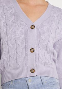 Světle levandulový pletený cardigan s kabelovým designem, výstřihem do V a třemi velkými hnědými knoflíky na přední straně, s ribbovaným lemem.