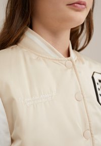 Šviesiai smėlio spalvos bomber tipo striukė su gipiūriniais apykaklėmis, mygtukais ir baltais rankovėmis. Dekoratyvinis siuvinėjimas skelbia: "Jei gali svajoti, gali tai padaryti."