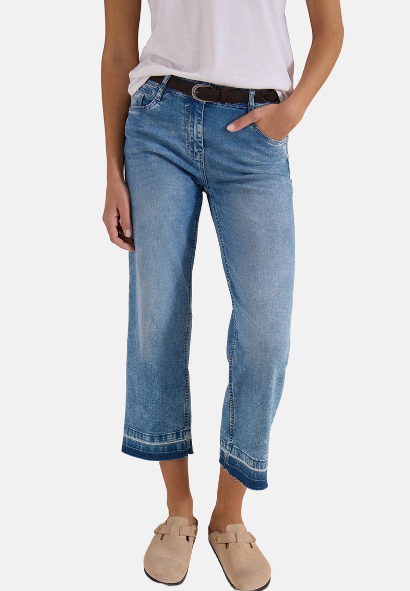 Femme portant un jean court bleu clair avec un ourlet foncé, un t-shirt blanc rentré, une ceinture noire et des chaussures beige à enfiler.