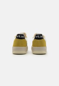 White sneaker with yellow suede accents and a black logo at the heel. Features a flat cream sole and fabric tongue. Simple and modern design.