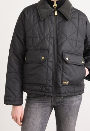 Personne portant une veste matelassée noire avec de grandes poches à rabat, des boutons-pression dorés, une fermeture éclair et un jean gris.