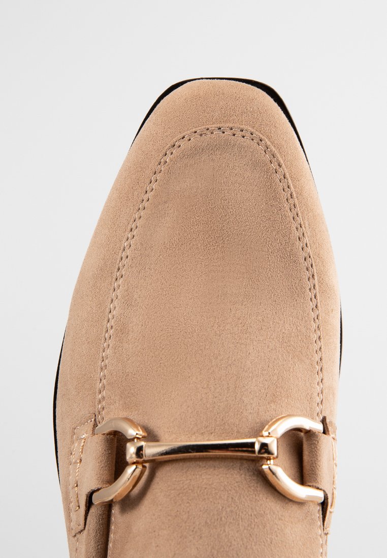 Beige Wildleder Slipper mit runder Zehenpartie, detaillierter Naht und einem goldfarbigen Metallelement über dem Rist. Glatte Oberfläche, zum Hineinschlüpfen.