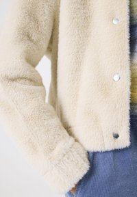 Cremfarbener, flauschiger Jacke mit Druckknöpfen, getragen über einem mehrfarbigen Pullover, kombiniert mit blauen Cordhosen, Hand in der Tasche.