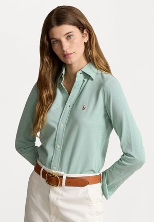 Camicia a colletto verde chiaro realizzata in un tessuto texturizzato, con bottoni e un piccolo logo ricamato. Abbinata a pantaloni bianchi e una cintura color beige.