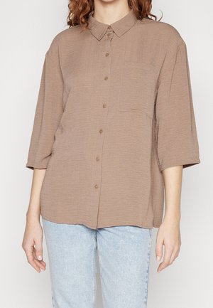 Femme portant une chemise marron ample boutonnée avec des manches trois-quarts et un jean bleu clair, vue du cou jusqu'aux hanches.