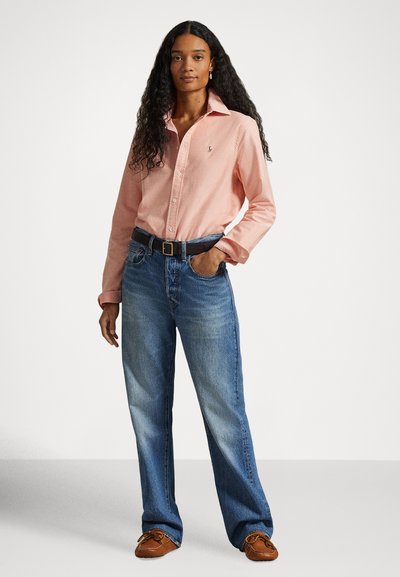 Gaiši rozā pogājama krekla krekls, taisnu kāju zili džinsi, brūnas mokasīnu stila kurpes. Krekls ar apkakli un izšūtu logotipu.