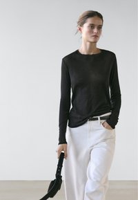 Woman with hair tied back wears black long-sleeve top, white pants with black belt, holding black bag strap in one hand, other hand in pocket.