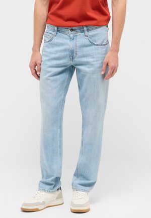 Mann trägt hellblaue Jeans mit geradem Bein, weiße Sneaker und ein rotes T-Shirt, steht vor einem schlichten hellen Hintergrund.