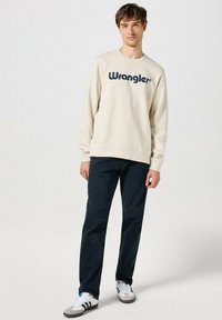 Felpa beige con logo "Wrangler" in navy, collo rotondo, maniche lunghe. Indossata con pantaloni blu scuro e sneakers bianche con stripature nere.