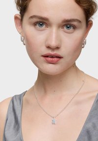 Collar de plata con un colgante en forma de oso, llevado sobre una blusa de satén gris. El modelo lleva pendientes de aro y maquillaje neutro.