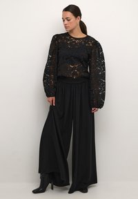 Haut en dentelle noire avec des manches longues bouffantes, superposé sur un pantalon large noir taille haute. Texture lisse, silhouette élégante, associé à des bottes noires.