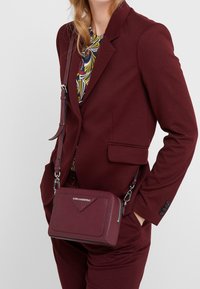 Bordowa torba na ramię od Karla Lagerfelda, prostokątna z teksturowanym wykończeniem, ze srebrnymi akcentami i przednim detalem w kształcie trójkąta. Regulowany pasek.