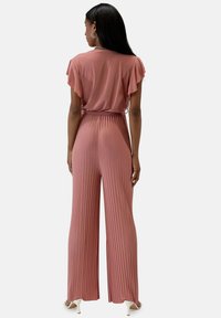 Rosa plisserad jumpsuit med korta volangärmar och knytning i midjan. Har vida byxor och ett mjukt, flödande tyg.