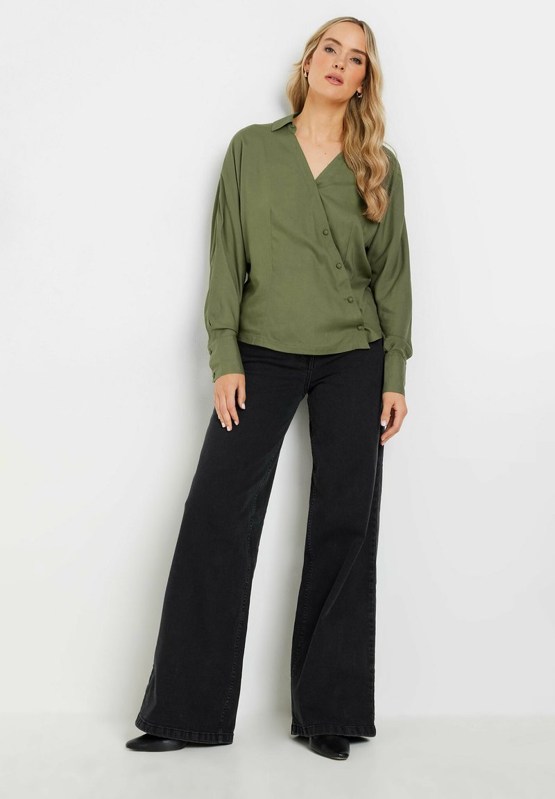 Blouse enveloppante vert olive à manches longues et détail de boutons, associée à un jean large noir et des bottines noires. Tissu lisse et léger.