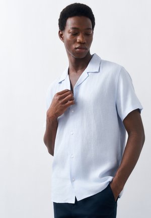 Νέος άνδρας με κοντά μαλλιά που φοράει ανοιχτό γαλάζιο πουκάμισο με κοντά μανίκια και κουμπιά, με το ένα χέρι να ρυθμίζει το γιακά του και το άλλο στο τσέπη.