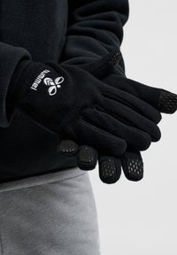 Svarta fleecehandskar med texturerat svart grepp på fingertopparna, med en vit logotyp på handleden. Mjukt material, ergonomisk design.