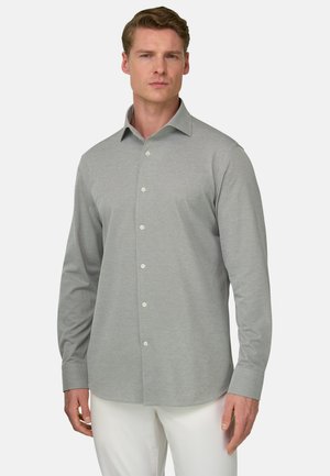 Homme portant une chemise à manches longues gris clair boutonnée et un pantalon blanc, debout devant un fond uni.