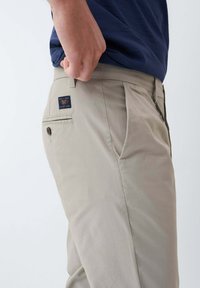 Světle béžové kalhoty s hladkým povrchem, mají boční kapsy a zadní kapsu s logem. Rovný střih, minimální detaily švů.