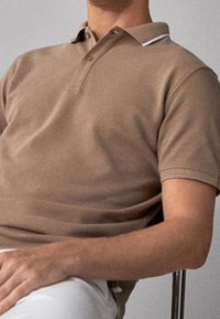 Koszulka polo z krótkim rękawem w beżowym kolorze z bawełny, z zapięciem na dwa guziki i białym akcentem na kołnierzyku. Noszona z białymi spodniami.