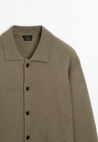 Cardigan en maille de laine marron avec col à revers et quatre boutons noirs sur le devant, posé à plat sur une surface blanche.