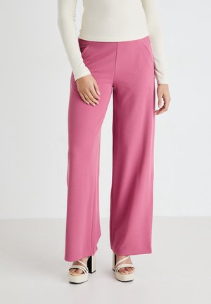 Mujer con pantalones anchos de talle alto de color rosa y tacones de plataforma blancos con tiras, de pie frente a un fondo blanco liso.