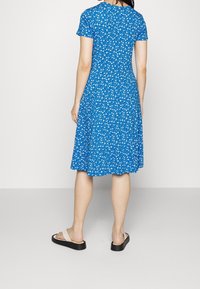 Femme debout portant une robe bleue à mi-longueur avec un imprimé floral blanc et des sandales blanches à semelles noires, de dos contre un fond uni.