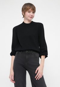 Black knitted sweater with a high neckline, puffed sleeves, and ribbed cuffs, paired with dark gray high-waisted jeans.