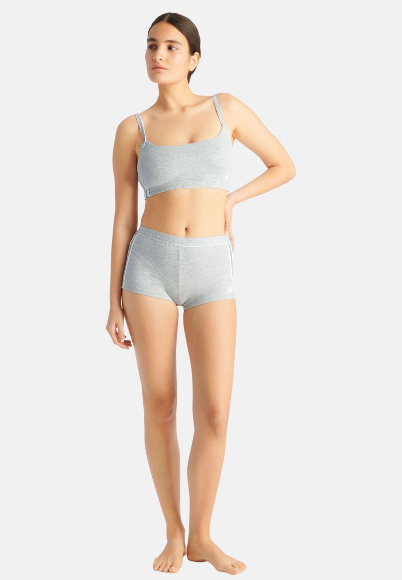 Donna in piedi a piedi nudi, che indossa un reggiseno sportivo grigio chiaro e pantaloncini abbinati, che guarda verso sinistra con una mano sul fianco, su sfondo bianco.