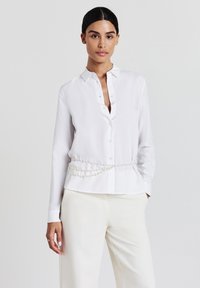 Donna che indossa una camicetta bianca con bottoni, una cintura di perle e pantaloni color crema, in piedi contro uno sfondo chiaro e semplice.