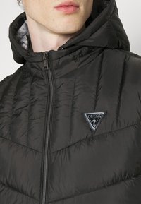 Schwarze Daunenjacke mit Kapuze, ausgestattet mit einem vertikalen Steppmuster und einem dreieckigen Guess-Logo auf der linken Brust. Sichtbarer Reißverschluss.