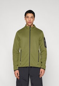 Φόρμα με φερμουάρ σε ελιά πράσινο χρώμα με ψηλό γιακά, υφή υφάσματος, τσέπες με μαύρο φερμουάρ και μαύρη λεπτομέρεια στο μανίκι.