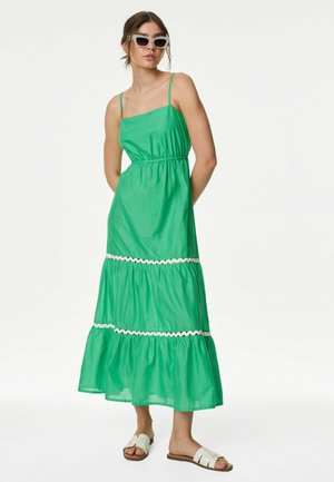 Grön bomullsmidi-klänning med spaghettiband, elastisk midja och vit spetskant på de lagerade ryscherna; kombinerad med vita sandaler och solglasögon.
