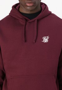 Burgundy huppari, joka on valmistettu puuvillasekoitteesta. Siinä on kengurutasku ja pieni valkoinen logo rinnassa. Mukana on myös nyörihuppu.